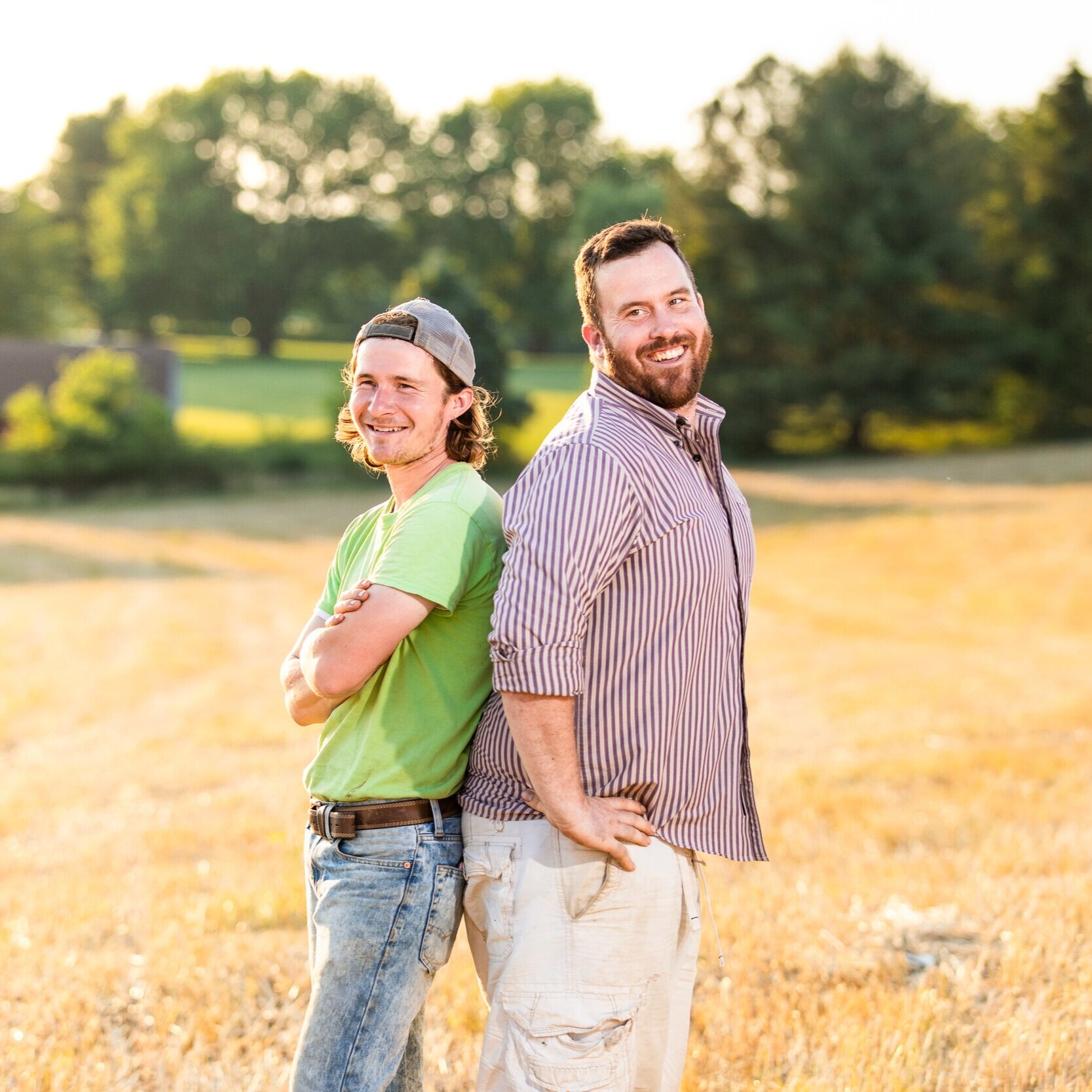 Two smiling men stand back-to-back in a sunlit field, showcasing a friendly camaraderie. One man wears a green t-shirt and a cap, while the other is in a striped button-up shirt. The background features lush trees and a golden landscape, emphasizing a warm, outdoor atmosphere.