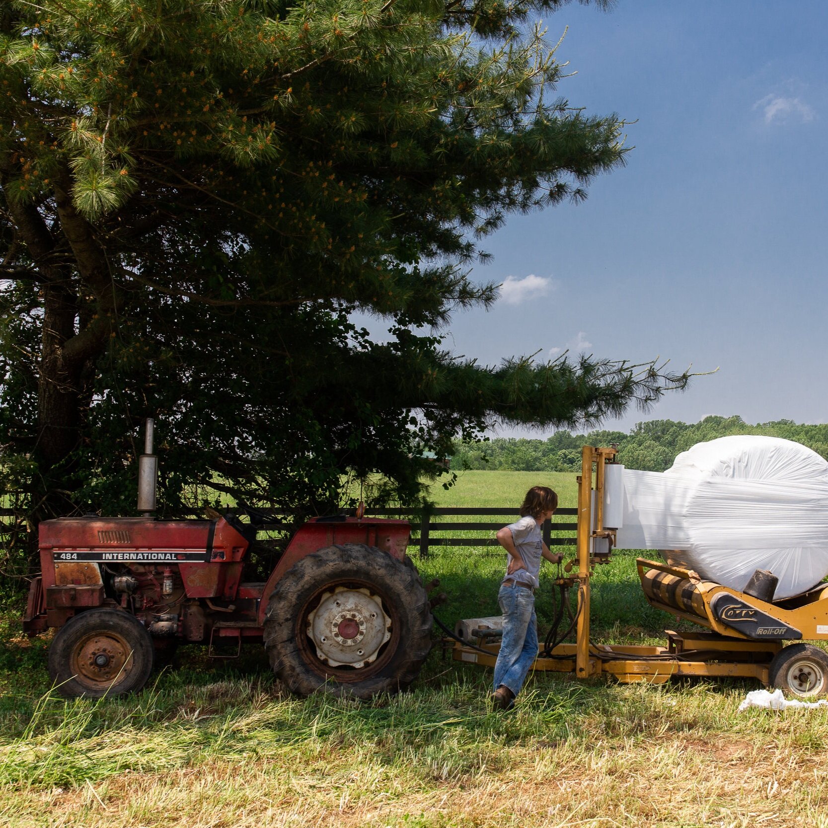 Farmer operating an International tractor next to a bale wrapper in a lush green field, surrounded by trees under a clear blue sky.