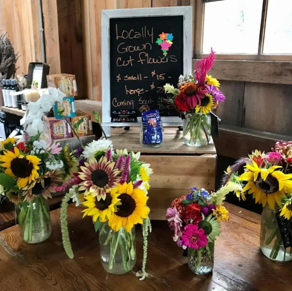 Locally grown cut flowers displayed in glass jars, featuring vibrant sunflowers, colorful blooms, and a chalkboard sign detailing pricing for small and large arrangements at a rustic market setting.