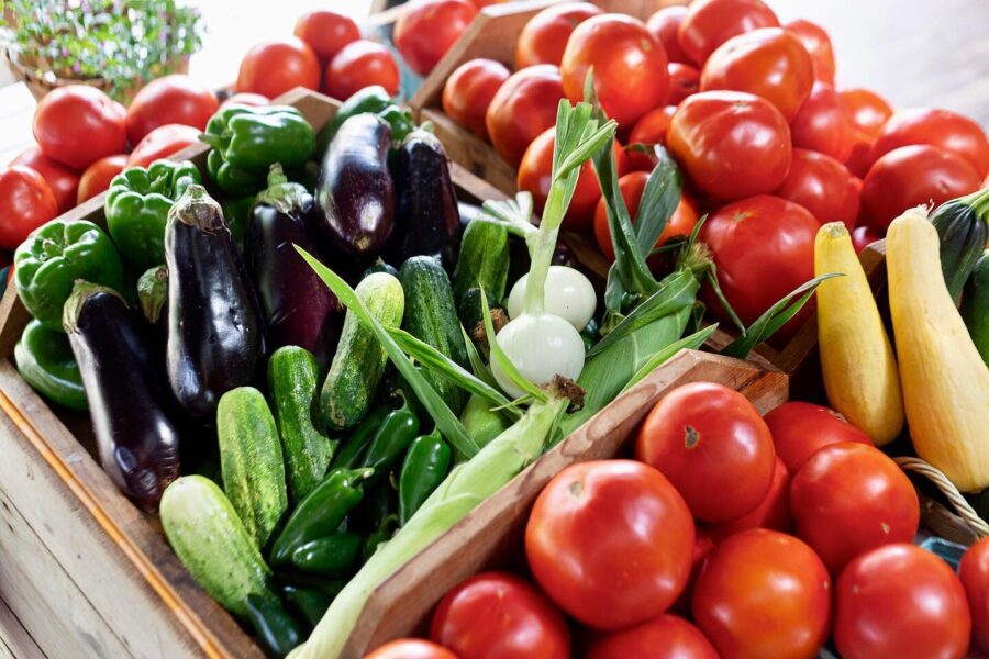 Colorful assortment of fresh vegetables including red tomatoes, green bell peppers, eggplants, cucumbers, yellow squash, and onions arranged in wooden crates at a farmers market.