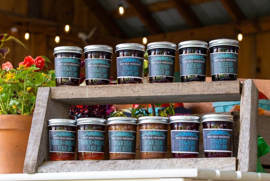Assorted jars of Neat Nick Preserves displayed on a wooden shelf, featuring flavors like Strawberry Balsamic Jam, Blueberry Ginger Jam, Bourbon Vanilla Peach Jam, Brown Sugar Apple Butter, and Strawberry Cherry Jam, with vibrant flowers in the background.