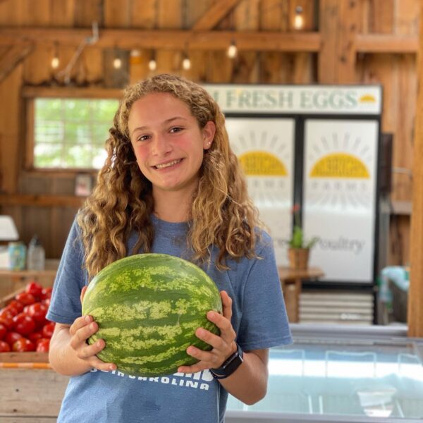 Girl smiling while holding a watermelon in a farm market setting, with fresh produce and a sign for eggs in the background.