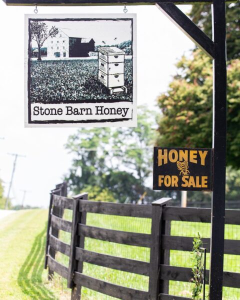 Sign for Stone Barn Honey featuring a beehive and farmhouse illustration, with an additional sign indicating honey for sale, set against a rural landscape with a wooden fence and green grass.