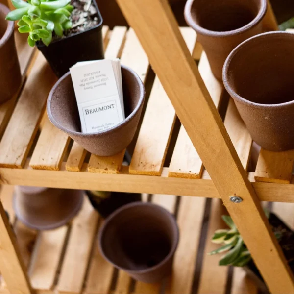 Wooden shelf displaying several clay pots and a small plant, with a business card for Beaumont Pottery placed inside one pot.