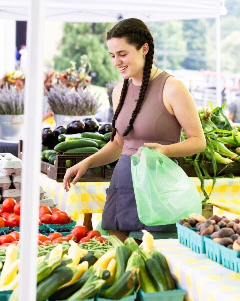 Young woman shopping for fresh vegetables at a farmers market, smiling while holding a green reusable bag. Colorful displays of tomatoes, zucchini, and other produce are arranged on tables under a canopy.