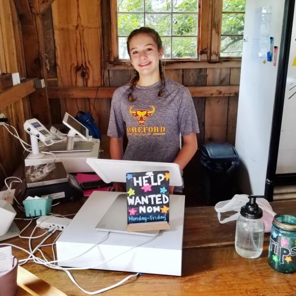 Smiling young girl at a rustic cash register in a wooden setting, wearing a grey shirt with a bull logo. A colorful sign reads 