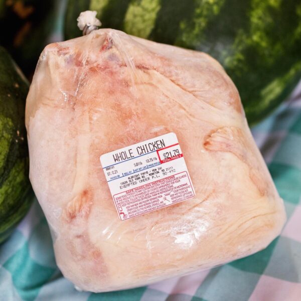 Whole chicken packaged in plastic with a price tag showing $1.79 per pound, surrounded by fresh produce on a checkered tablecloth.